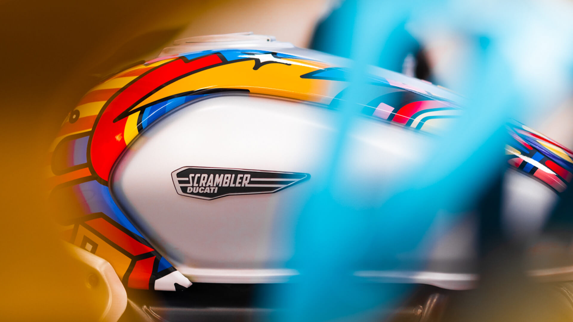 dettaglio del serbatoio con il logo di una scrambler ducati su una moto con una livrea multicolore