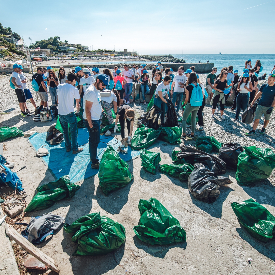 persone impegnate in un'attività di pulizia di una spiaggia, raccogliendo rifiuti in numerosi sacchi neri e verdi